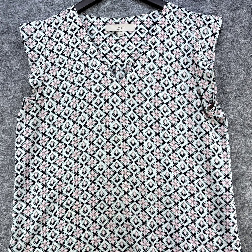 A sleeveless V-neck top with a geometric diamond pattern in black, white, and light pink, hanging on a black hanger against a gray textured background.