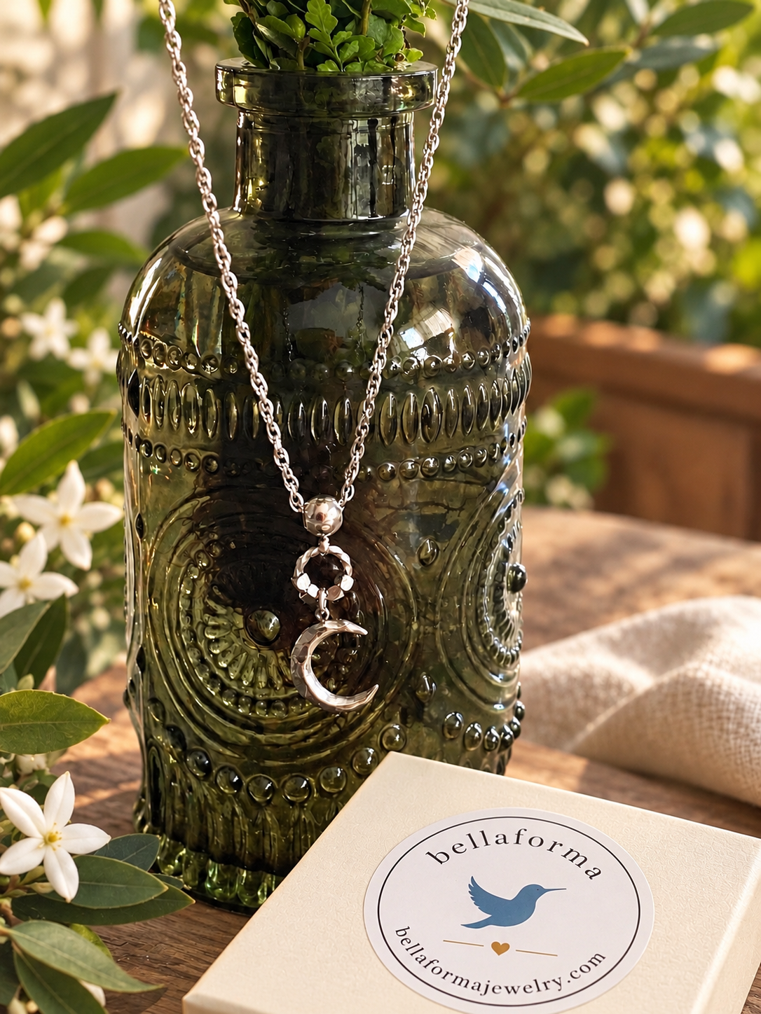 A silver crescent moon pendant necklace with a beaded ring detail, draped over a dark green glass bottle. The bottle is set on a wooden table with green foliage and white flowers in the background, next to a cream-colored jewelry box with a 'bellaforma' brand label.