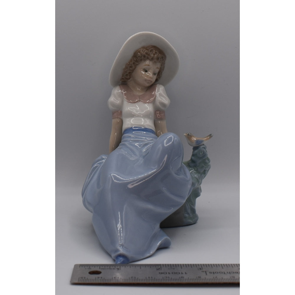 Width view of Lladro porcelain figurine of a woman with a white hat and blue dress, sitting on a rock, with a ruler for scale.