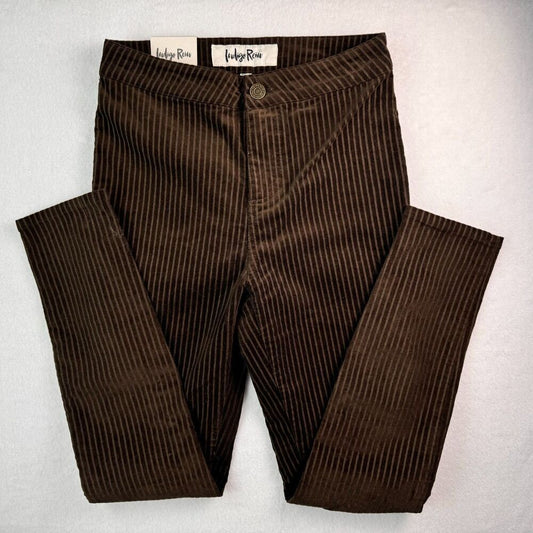 A pair of dark brown pinstriped corduroy pants laid flat on a light gray background, featuring a button closure and visible 'Indigo Room' brand labels at the waistband.