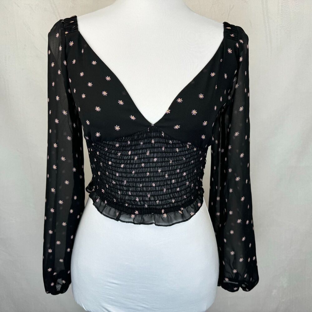A black long-sleeve blouse with a smocked bodice and delicate pink floral embroidery, displayed on a white mannequin torso against a light gray fabric backdrop.
