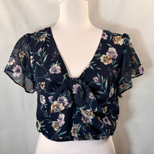 A navy blue short-sleeve blouse with a floral print, displayed on a white mannequin torso. The blouse features a deep V-neckline, a front tie detail, and sheer flutter sleeves.
