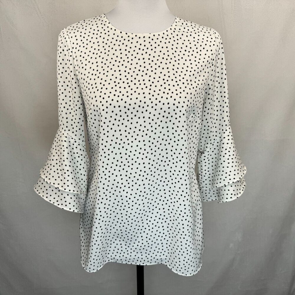 A white long-sleeve blouse with a black polka dot pattern, displayed on a mannequin against a light gray fabric backdrop.