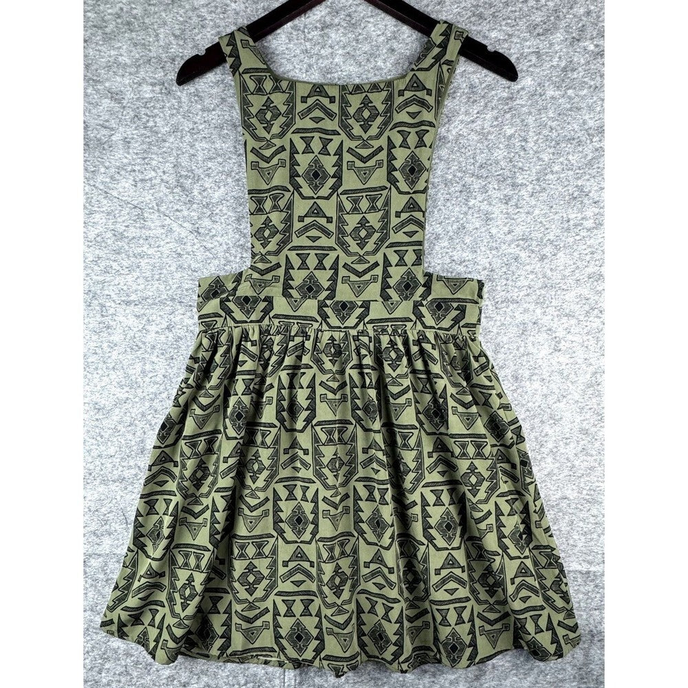 A sleeveless children's dress with a geometric tribal-style pattern in olive green and black, hanging on a black hanger against a textured gray background.