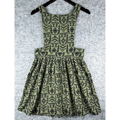 A sleeveless children's dress with a geometric tribal-style pattern in olive green and black, hanging on a black hanger against a textured gray background.