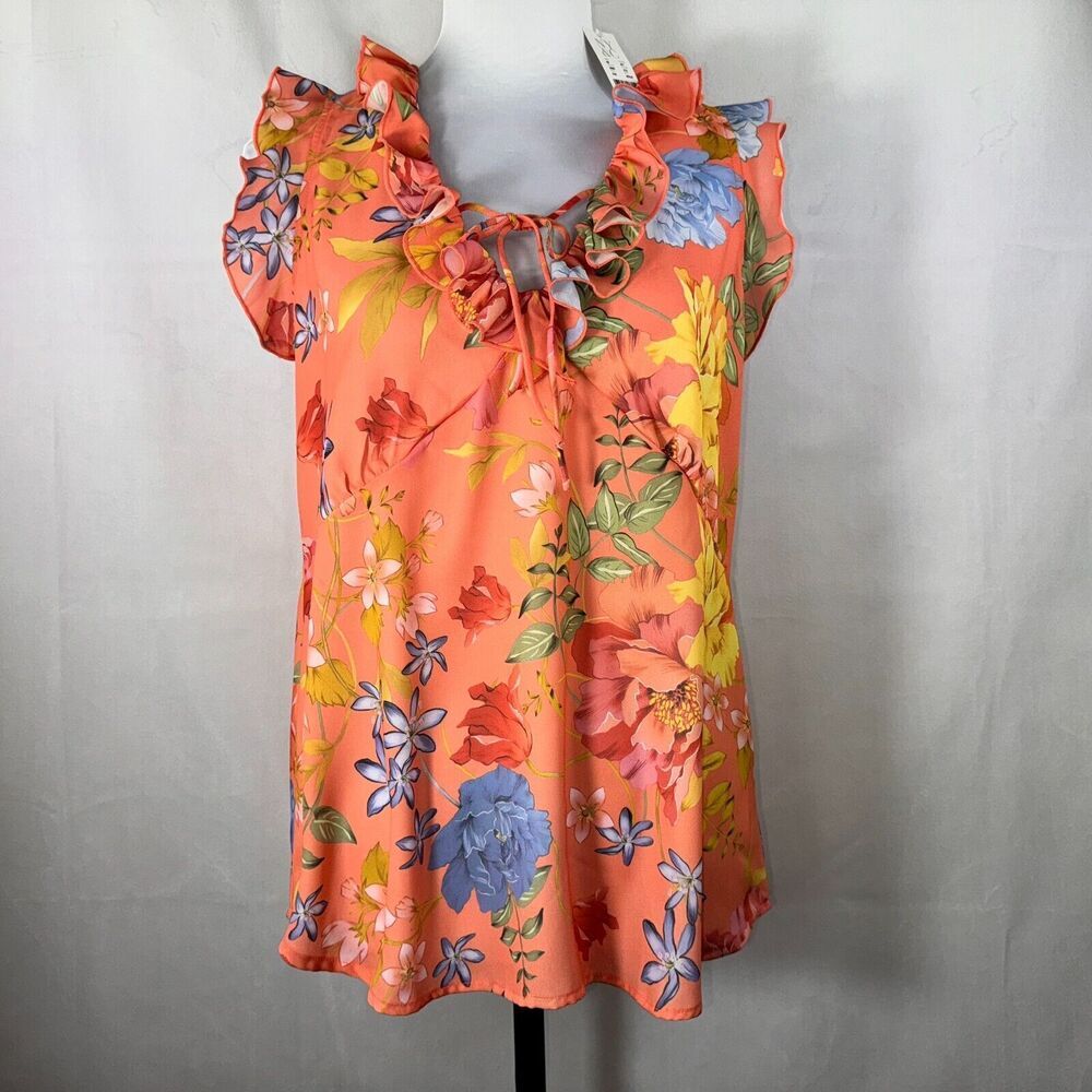 A women's sleeveless blouse with ruffled shoulders and a drawstring neckline, featuring a vibrant orange base with a multicolor floral print in red, yellow, blue, and green, displayed on a mannequin against a light gray backdrop.