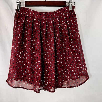 A dark red skirt with a small white and black paisley print, hanging on a wooden hanger against a white fabric background.