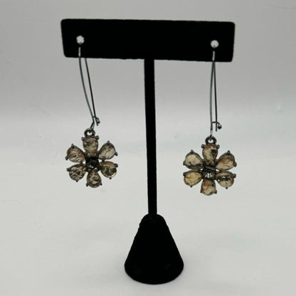 A pair of dangling flower-shaped earrings displayed on a black T-stand against a light gray background. The earrings have clear faceted stones set in a dark metal with a gold-toned center.