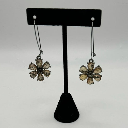 A pair of dangling flower-shaped earrings displayed on a black T-stand against a light gray background. The earrings have clear faceted stones set in a dark metal with a gold-toned center.