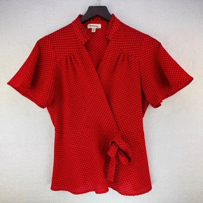 A red short-sleeve wrap blouse with a small white polka dot pattern, featuring a collar, front wrap closure, and a tie detail at the waist, hanging on a black hanger against a light gray textured wall.