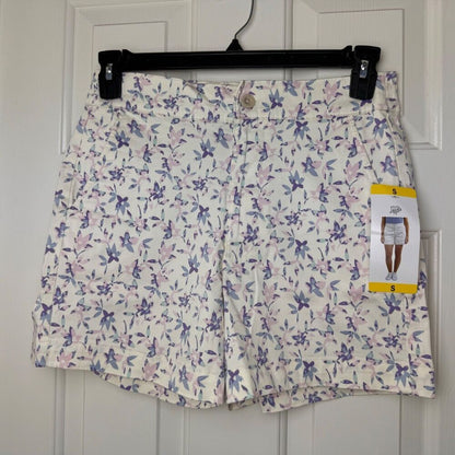 A pair of women's floral print shorts in white, featuring purple and pink flowers, hanging on a black hanger against a white paneled door. A yellow and white size tag is attached to the right side.