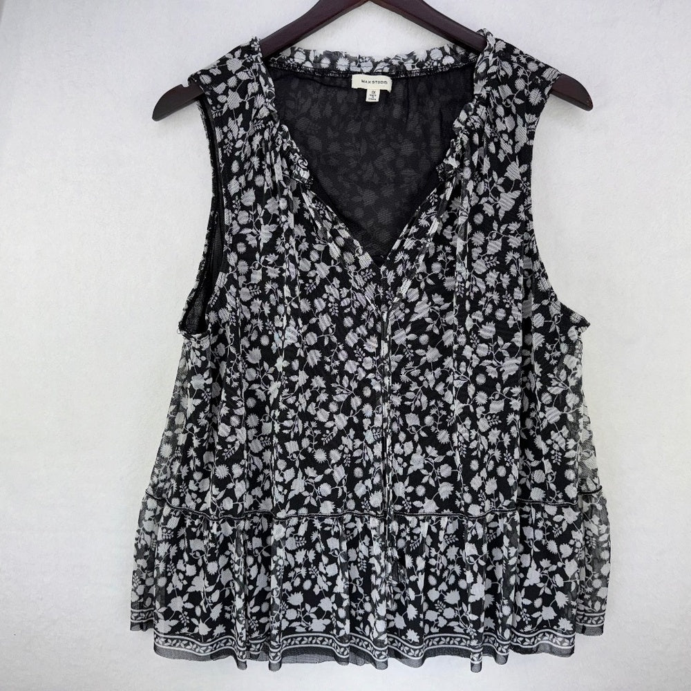 A sleeveless black and white floral print blouse hanging on a dark wooden hanger against a light gray wall. The blouse features a V-neckline, tiered ruffle hem, and a small white brand label at the inner collar.