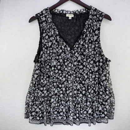 A sleeveless black and white floral print blouse hanging on a dark wooden hanger against a light gray wall. The blouse features a V-neckline, tiered ruffle hem, and a small white brand label at the inner collar.
