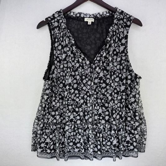 A sleeveless black and white floral print blouse hanging on a dark wooden hanger against a light gray wall. The blouse features a V-neckline, tiered ruffle hem, and a small white brand label at the inner collar.