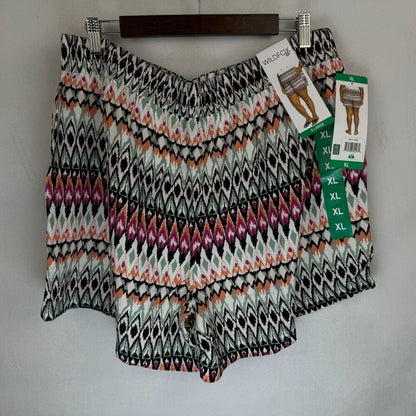A pair of women's shorts with an elastic waistband and a colorful geometric pattern, hanging on a wooden hanger against a light gray wall. The shorts feature a multicolor diamond and zigzag pattern in black, white, pink, orange, and green.