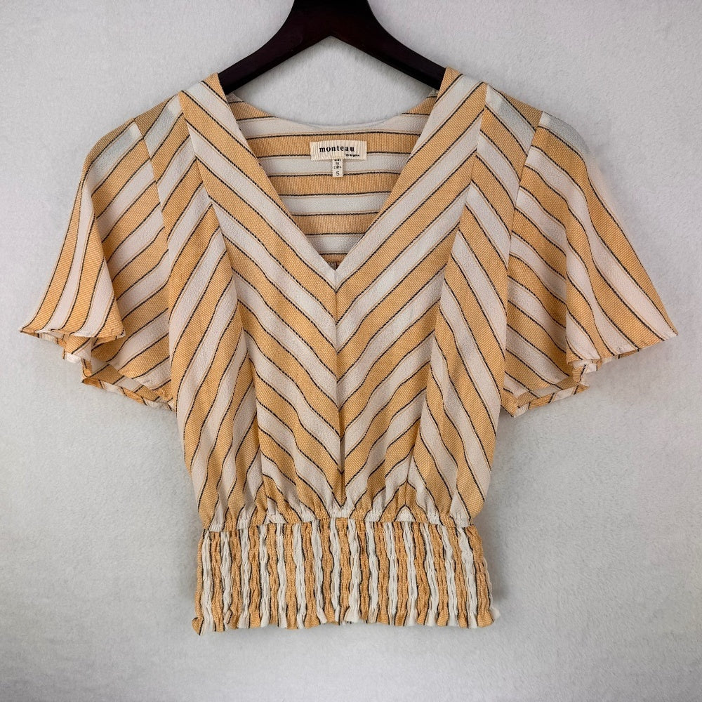 A short-sleeve V-neck blouse with a chevron stripe pattern in mustard yellow and off-white, hanging on a dark wooden hanger against a textured light gray wall. The brand name 'Montoya' is visible on the inner neck label.