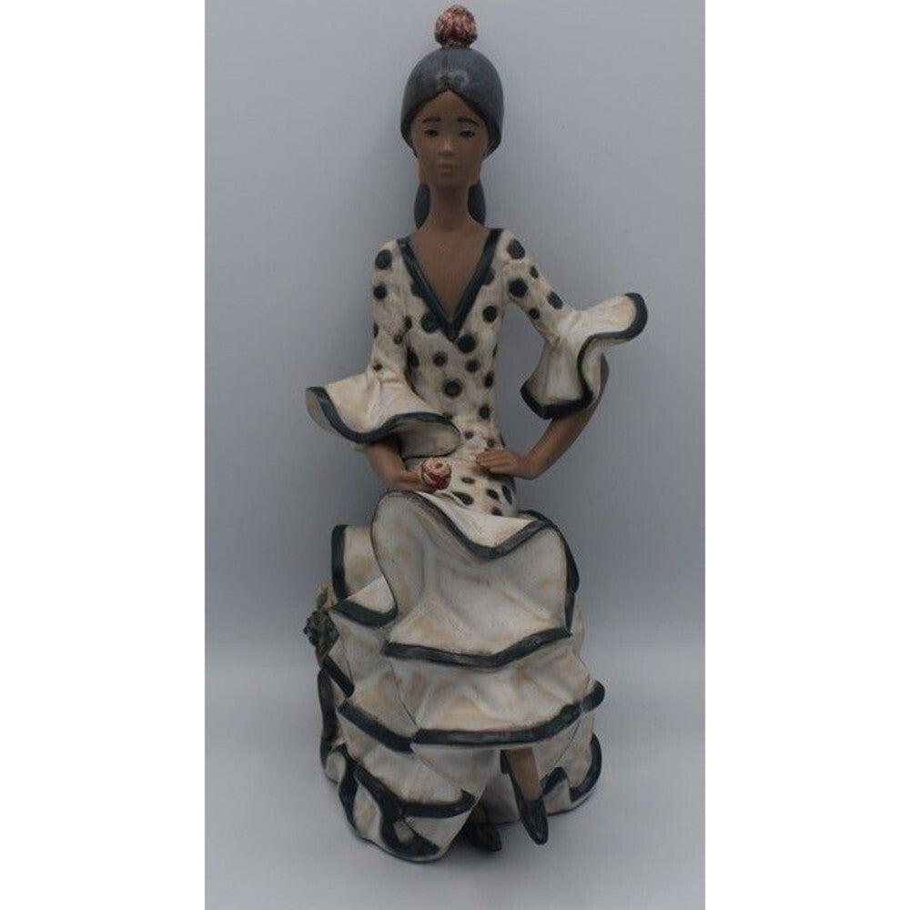 A ceramic figurine of a seated woman in a flamenco-style dress with black polka dots and black trim, set against a plain gray background.