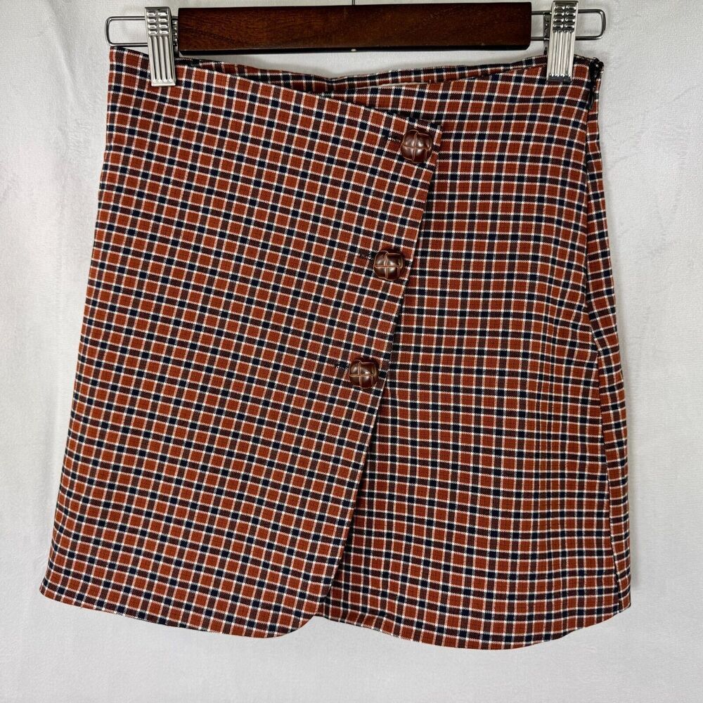 A plaid wrap skirt in a rust red, black, and white pattern, hanging on a wooden hanger with metal clips against a white fabric background.