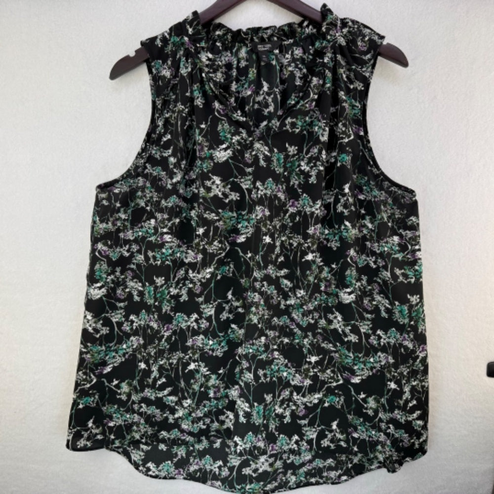 A sleeveless black blouse with a dense green, white, and purple floral print, hanging on a dark wooden hanger against a textured off-white wall.