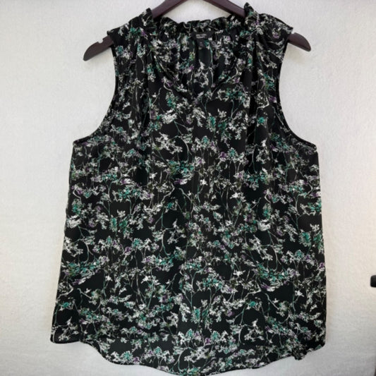 A sleeveless black blouse with a dense green, white, and purple floral print, hanging on a dark wooden hanger against a textured off-white wall.