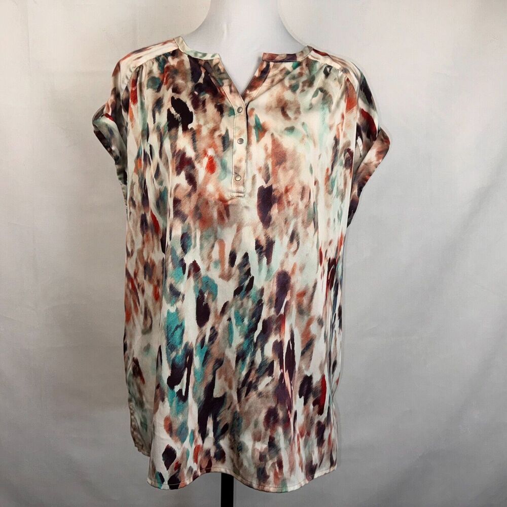 A short-sleeve button-front blouse with an abstract watercolor-style print in beige, brown, teal, and burgundy, displayed on a white mannequin torso against a light gray fabric backdrop.