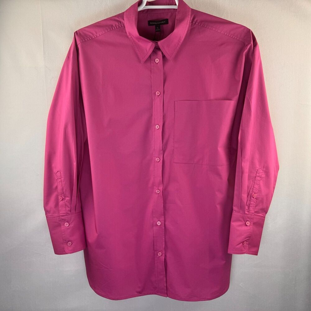 A bright pink long-sleeve button-down shirt with a single chest pocket, hanging on a white hanger against a light gray fabric backdrop.