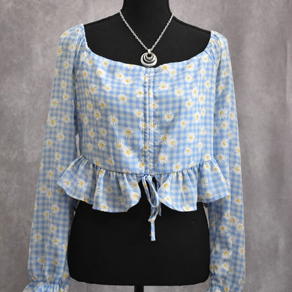 A light blue and white gingham floral long-sleeve blouse displayed on a black dress form mannequin, featuring a square neckline, ruffled hem, and front tie detail.