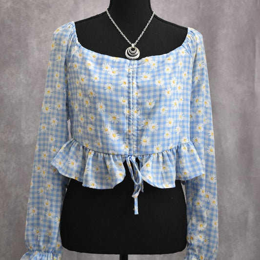 A light blue and white gingham floral long-sleeve blouse displayed on a black dress form mannequin, featuring a square neckline, ruffled hem, and front tie detail.