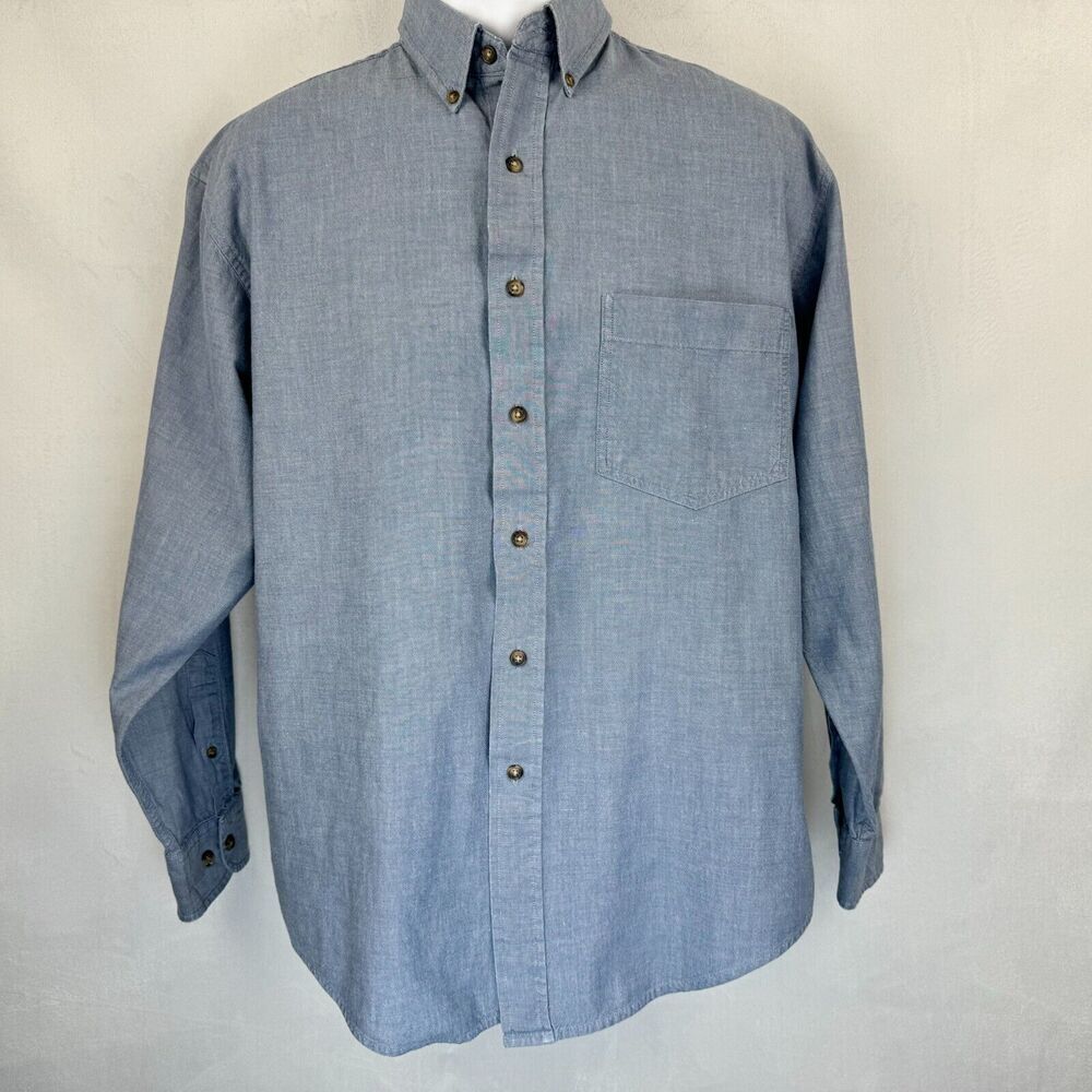 A light blue chambray long-sleeve button-down shirt with a single chest pocket and brown buttons, displayed on a mannequin torso against a light gray textured background.