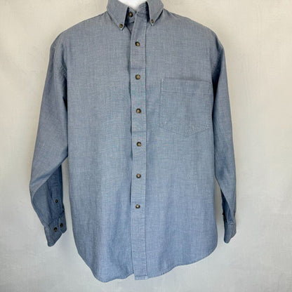 A light blue chambray long-sleeve button-down shirt with a single chest pocket and brown buttons, displayed on a mannequin torso against a light gray textured background.