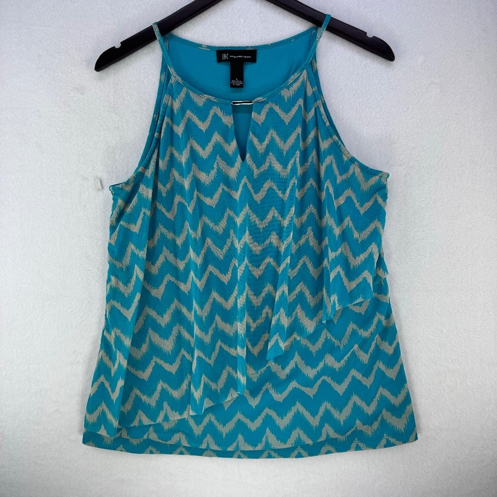 A sleeveless tank top with a turquoise and beige ikat-style chevron pattern, hanging on a black hanger against a textured white wall.