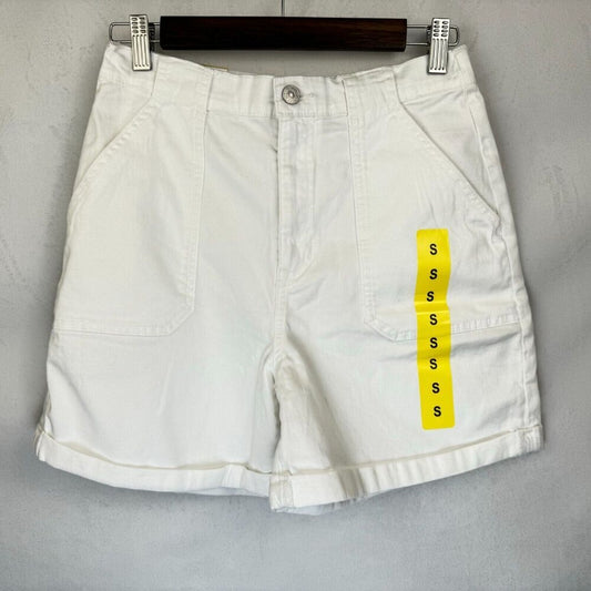 White denim shorts with a yellow size tag hanging from the side pocket, displayed on a dark wooden hanger against a gray textured wall.