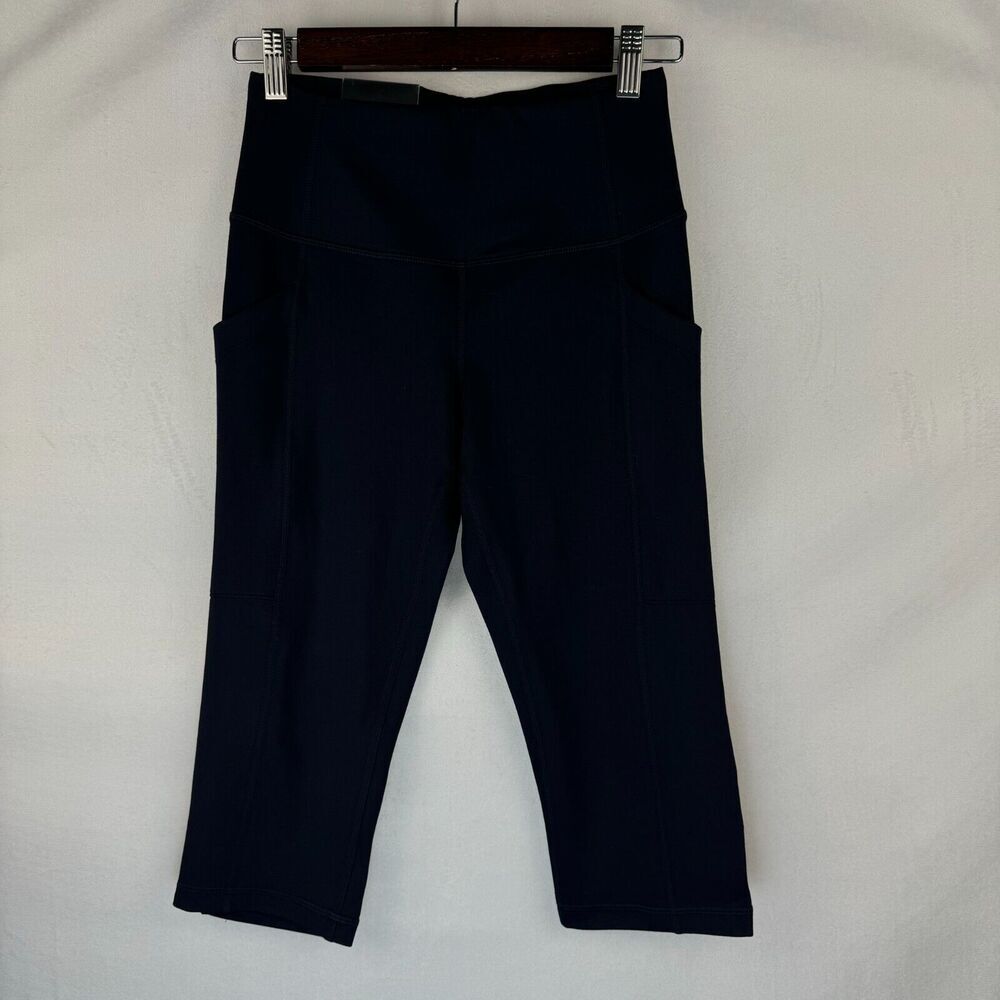 Dark navy blue high-waisted activewear capri pants with side pockets, hanging from a wooden hanger with metal clips against a white fabric background.