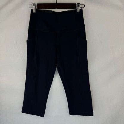 Dark navy blue high-waisted activewear capri pants with side pockets, hanging from a wooden hanger with metal clips against a white fabric background.