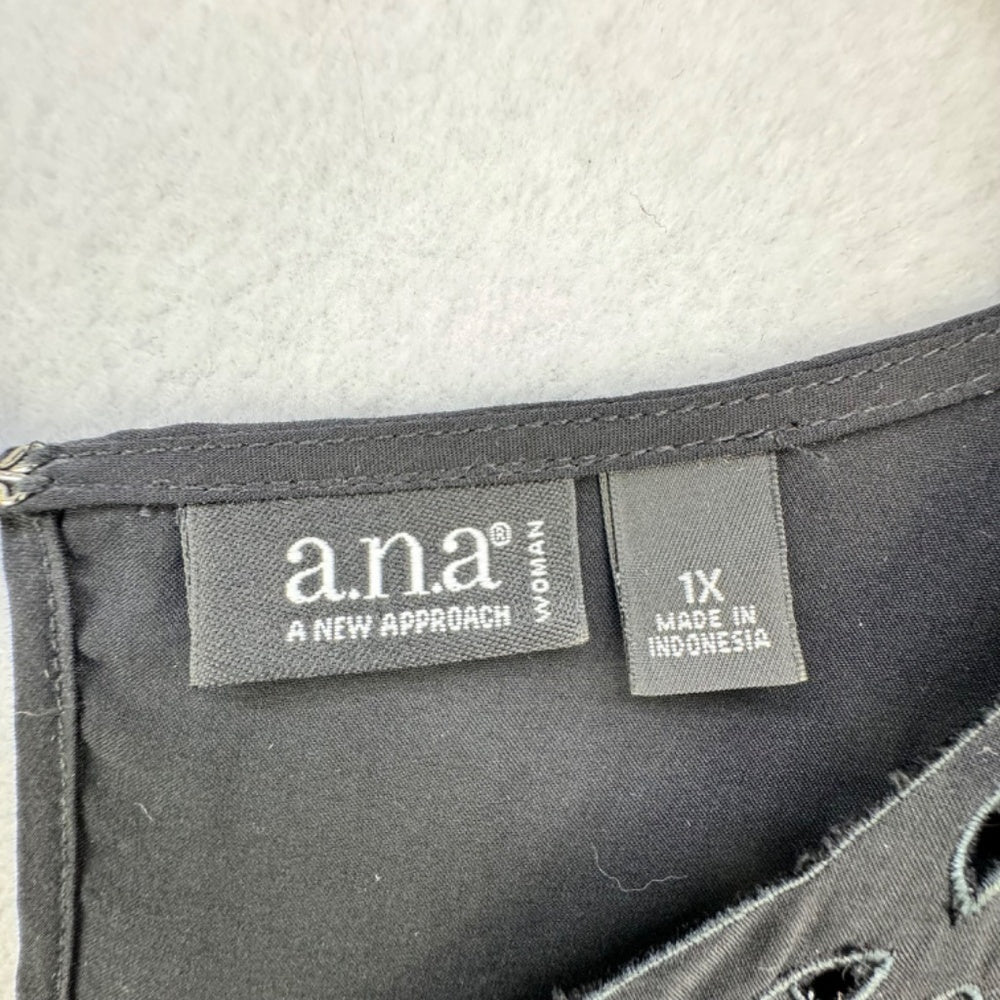 Close-up of a black garment's interior label area showing the 'ana A NEW APPROACH' brand tag and a size '1X MADE IN INDONESIA' tag on a light gray fabric background.