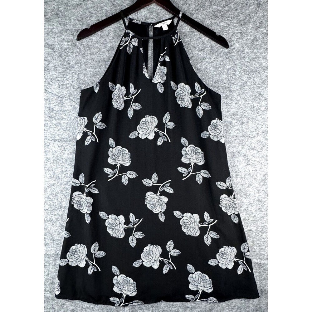 A black halter-neck dress with a white rose and leaf floral pattern, hanging on a dark wooden hanger against a textured gray background.