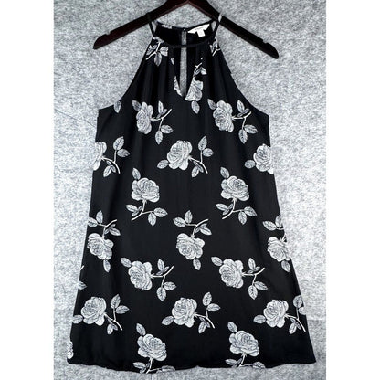 A black halter-neck dress with a white rose and leaf floral pattern, hanging on a dark wooden hanger against a textured gray background.
