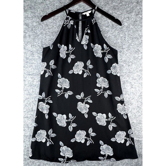 A black halter-neck dress with a white rose and leaf floral pattern, hanging on a dark wooden hanger against a textured gray background.
