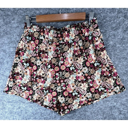 A pair of women's floral shorts with a dense paisley and daisy pattern, hanging on a wooden hanger against a gray textured background.