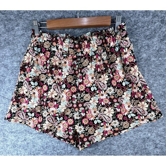 A pair of women's floral shorts with a dense paisley and daisy pattern, hanging on a wooden hanger against a gray textured background.