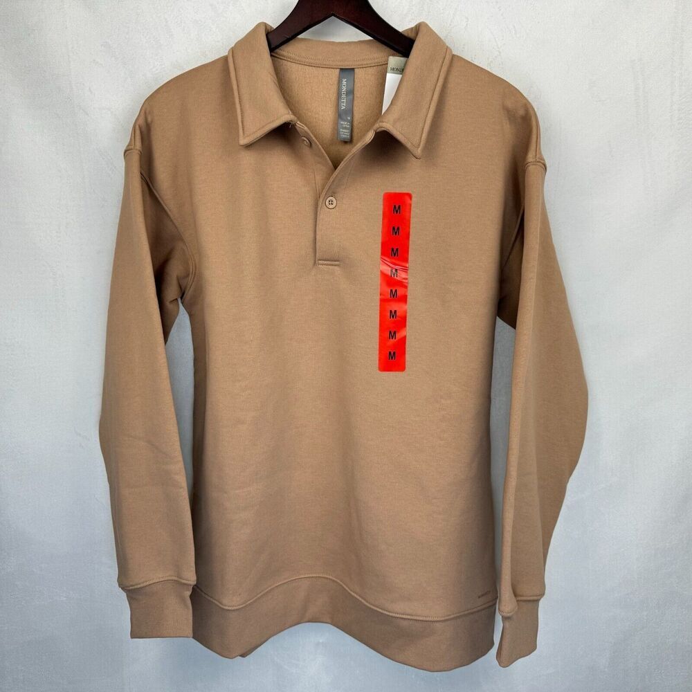 A tan long-sleeve polo shirt hanging on a dark wooden hanger against a light gray textured wall. The shirt features a vertical red paper tag with black text on the left chest.