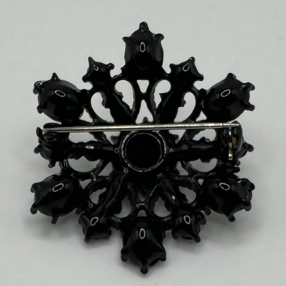 A vintage-style black enamel brooch featuring a central circular opening surrounded by eight ornate, petal-like extensions, with a silver-tone metal pin bar visible across the middle, set against a plain light gray background.