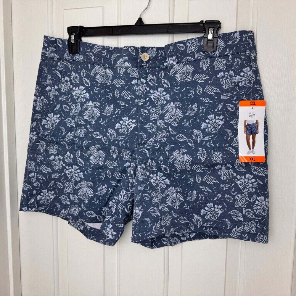 A pair of navy blue shorts with a white tropical floral and leaf pattern, hanging on a black hanger against a white paneled door. A brand tag is attached to the right leg.