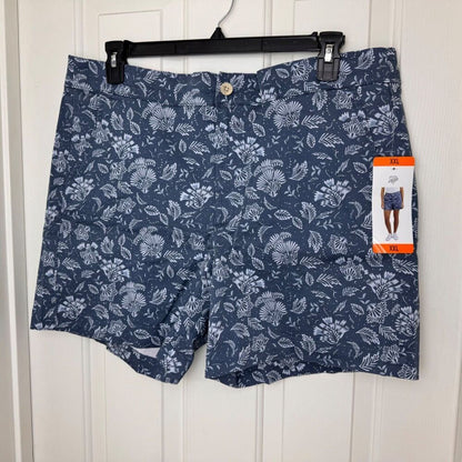 A pair of navy blue shorts with a white tropical floral and leaf pattern, hanging on a black hanger against a white paneled door. A brand tag is attached to the right leg.