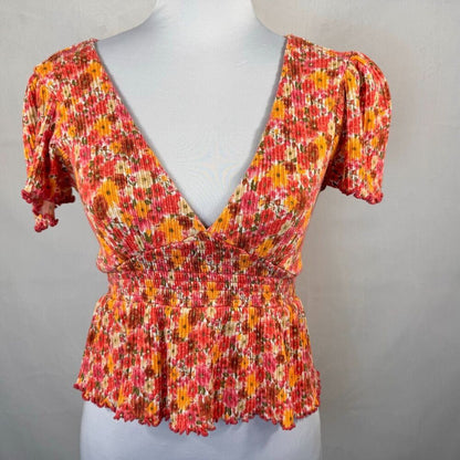 A short-sleeve, deep V-neck blouse with a smocked waist and ruffled hem, featuring a vibrant red, orange, and yellow floral pattern, displayed on a white mannequin torso.