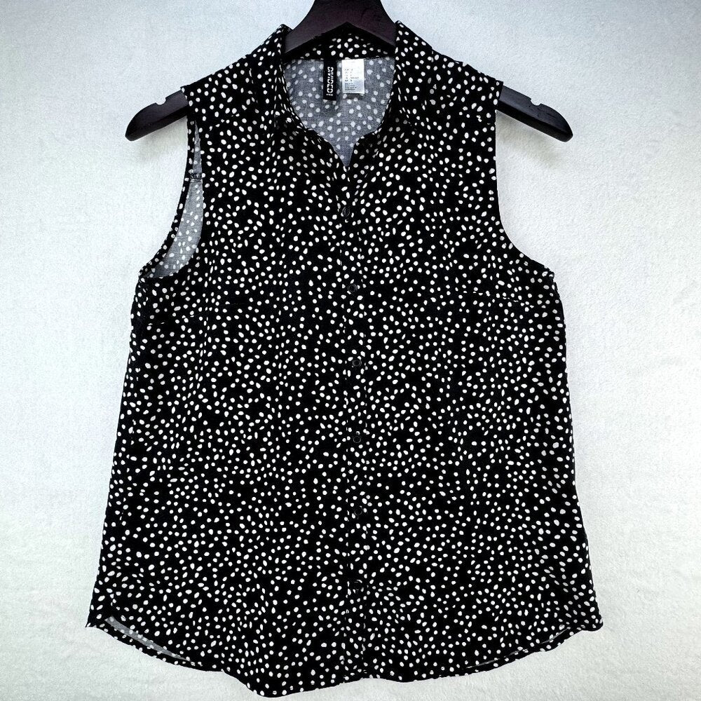 A sleeveless black blouse with a white micro-dot pattern, featuring a pointed collar and button-down front, hanging on a black hanger against a textured white background.