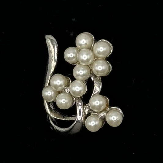 A silver-toned ring featuring a cluster of white faux pearls arranged in floral and vine-like patterns, set against a solid black background.