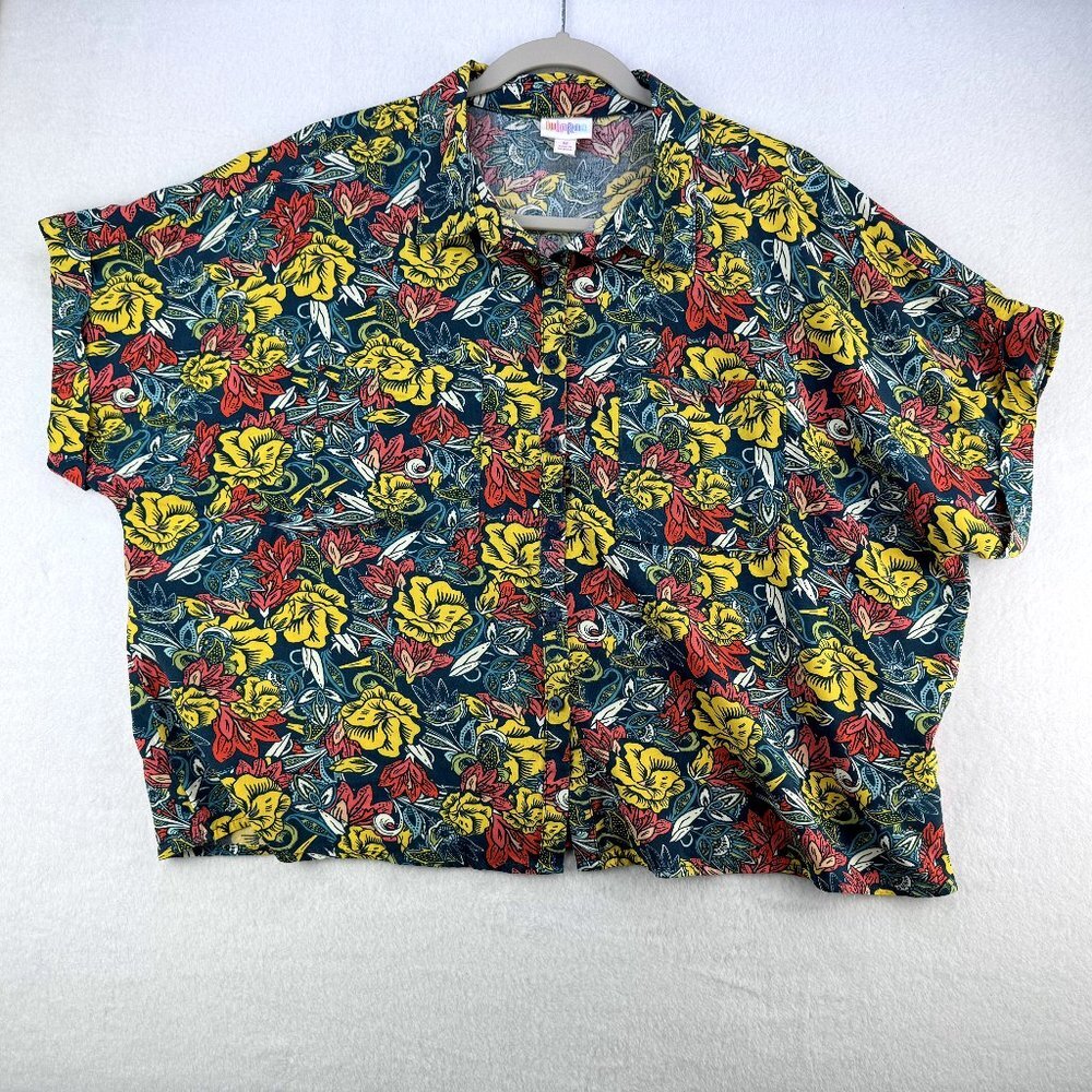 A short-sleeve button-up shirt with a dense yellow, red, and green floral print on a dark background, hanging on a gray hanger against a white textured wall.