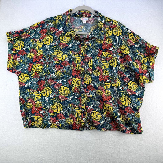 A short-sleeve button-up shirt with a dense yellow, red, and green floral print on a dark background, hanging on a gray hanger against a white textured wall.
