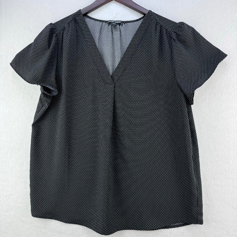 A black short-sleeve blouse with a subtle white polka dot pattern, featuring a V-neckline and puff sleeves, hanging on a dark wooden hanger against a light gray textured wall.